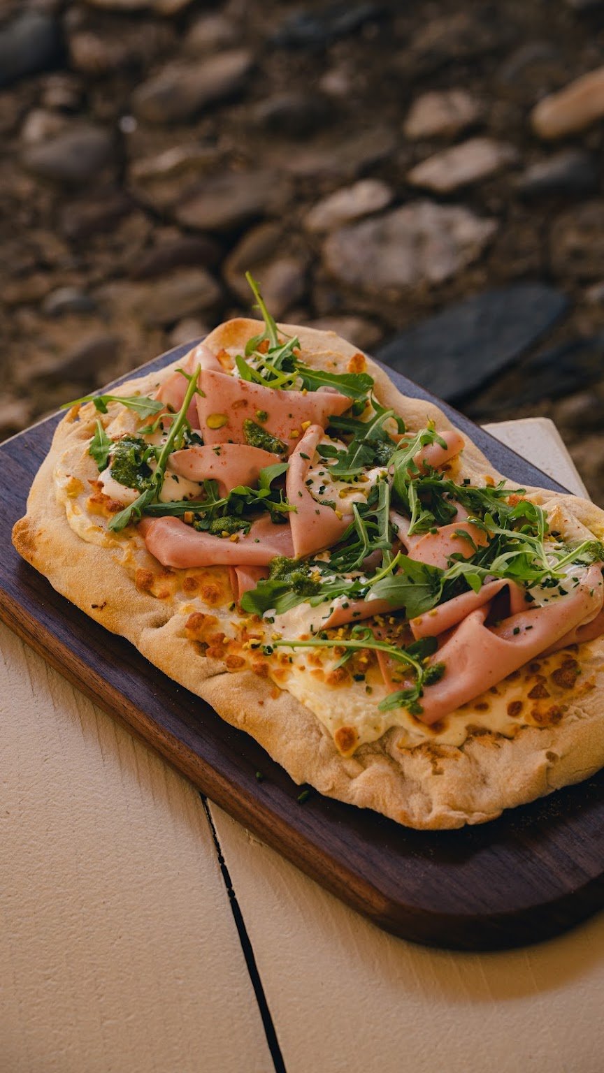 Focaccia garnie de fines tranches de jambon italien, roquette et noisettes, sur une planche en bois.