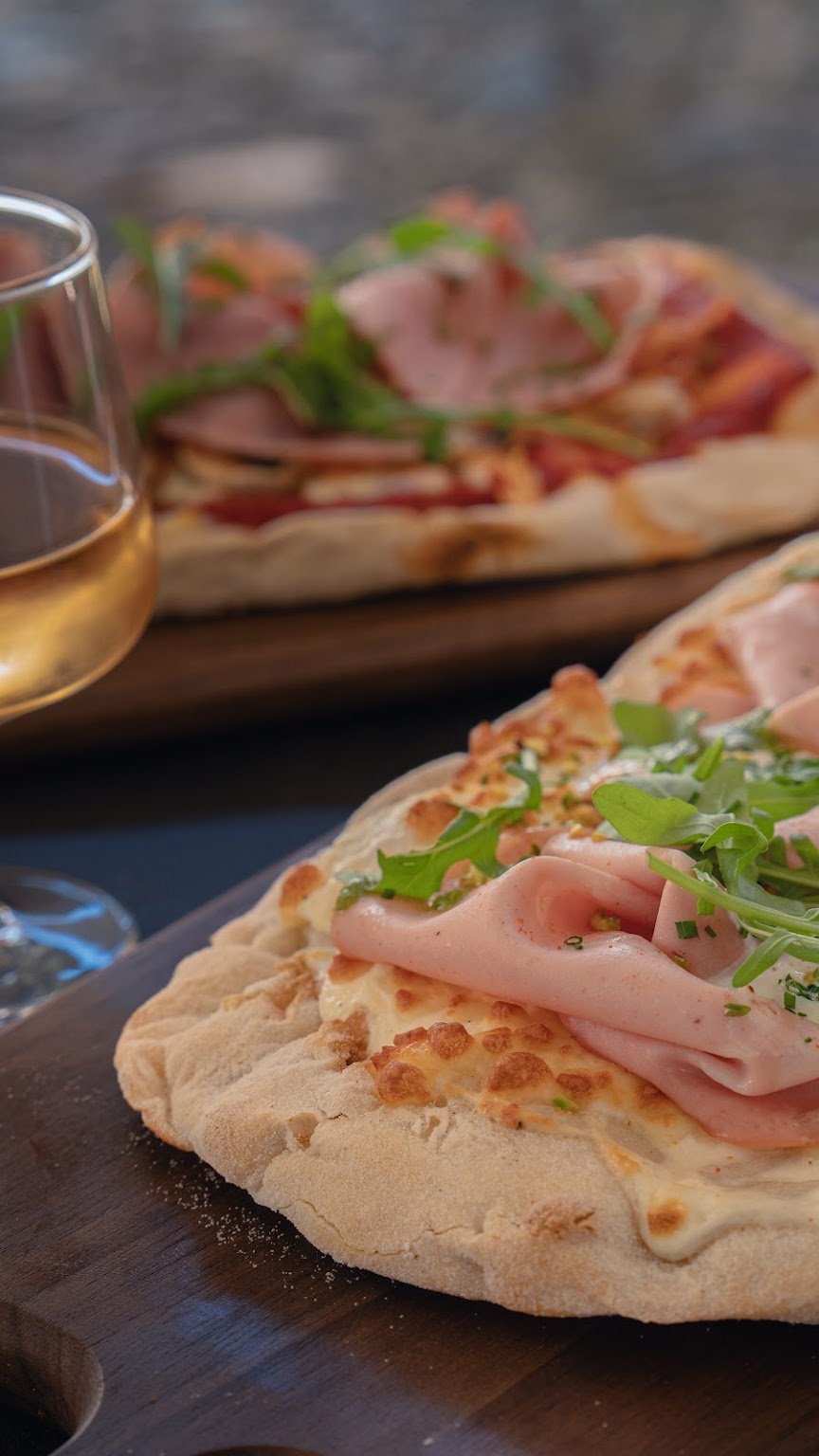 Pinsa au jambon et à la roquette servie sur une planche, un verre de vin blanc à côté.