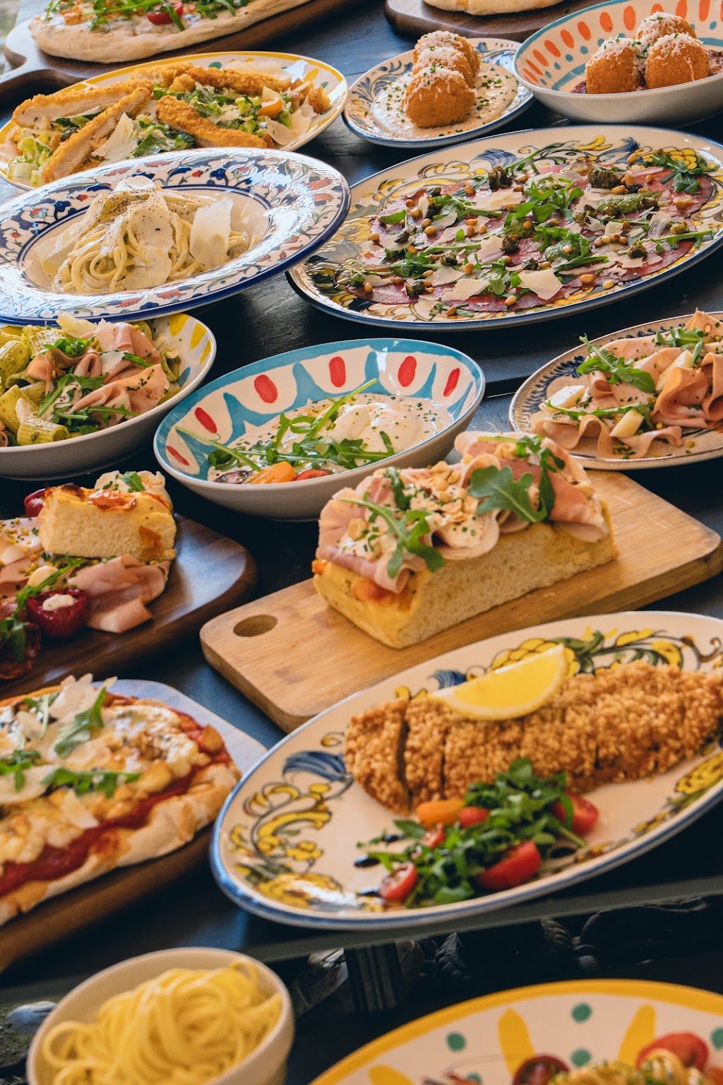 Une grande tablée : pinsas, pâtes, focaccias et arancini servis dans des assiettes en céramique peinte, toutes différentes.