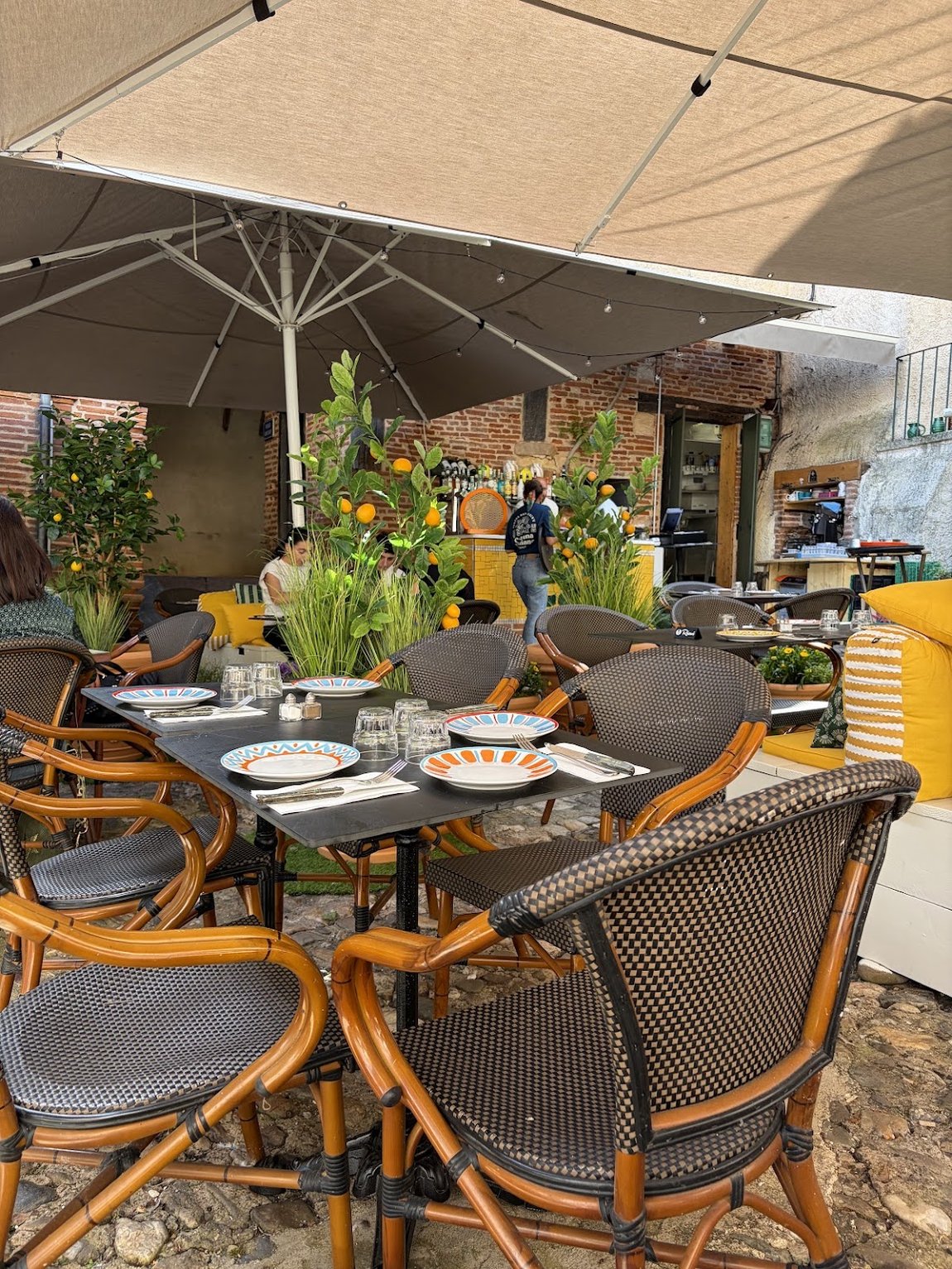 La terrasse de Cali : parasols, citronniers en pot, chaises en rotin et tables dressées d’assiettes en céramique peinte, devant un mur de brique.